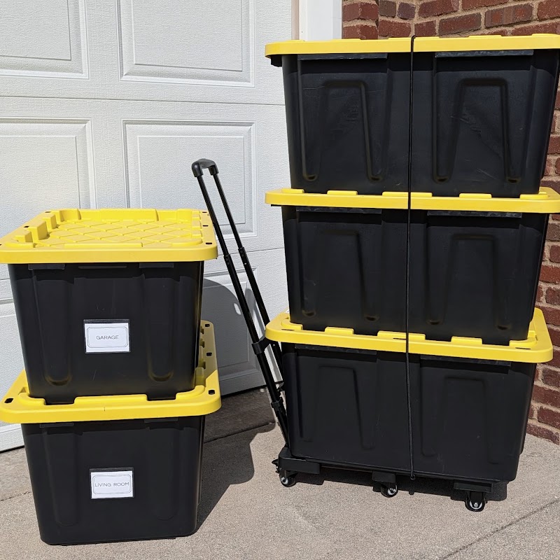 Twenty heavy-duty black moving totes with yellow lids, labeled Garage and Living Room, stacked next to a folding dolly by a garage door