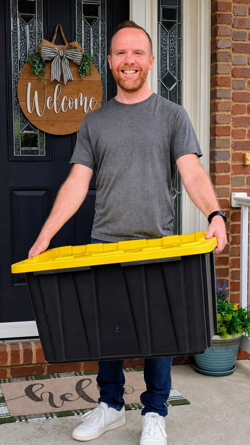 Ryan, owner of Crate & Key, smiling on a front porch and holding a black moving tote with a yellow lid